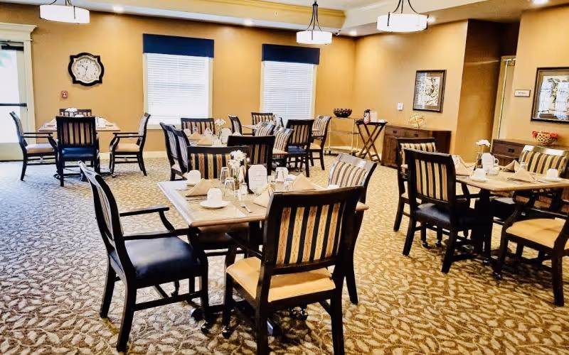 Dining room with multiple tables set for meals and wooden chairs in a carpeted, well-lit room.