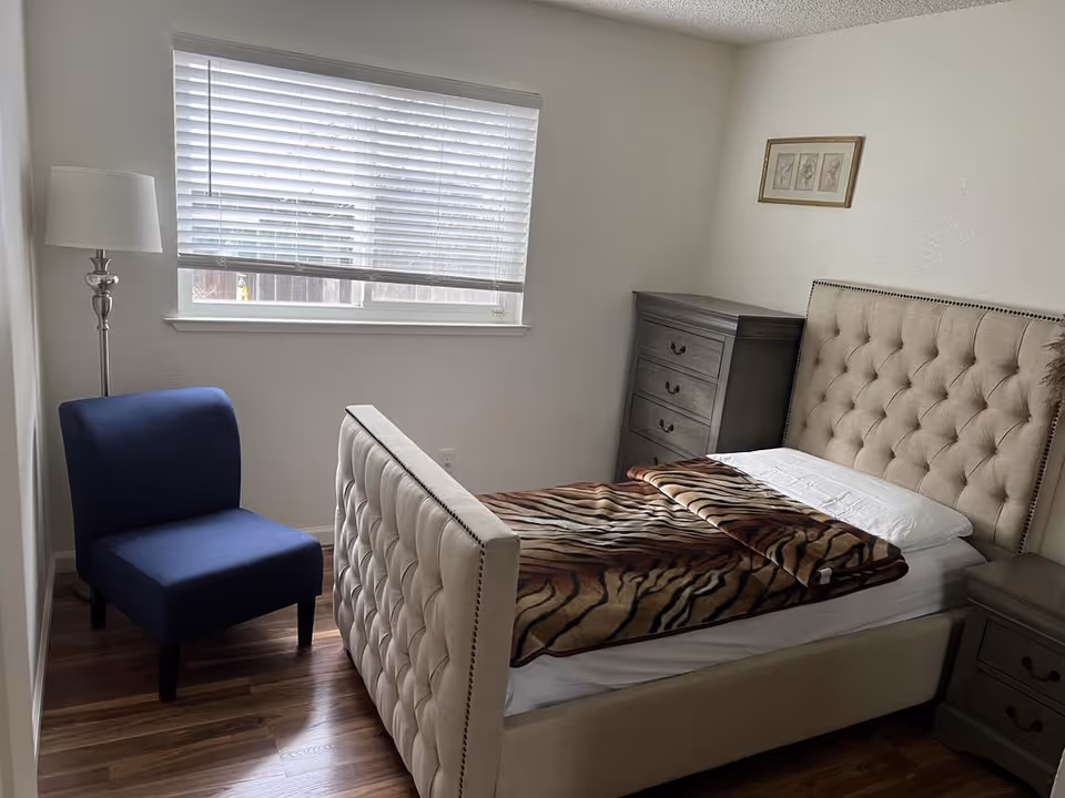 A furnished senior living bedroom featuring a tufted headboard, dresser, window, blue chair, and zebra blanket.
