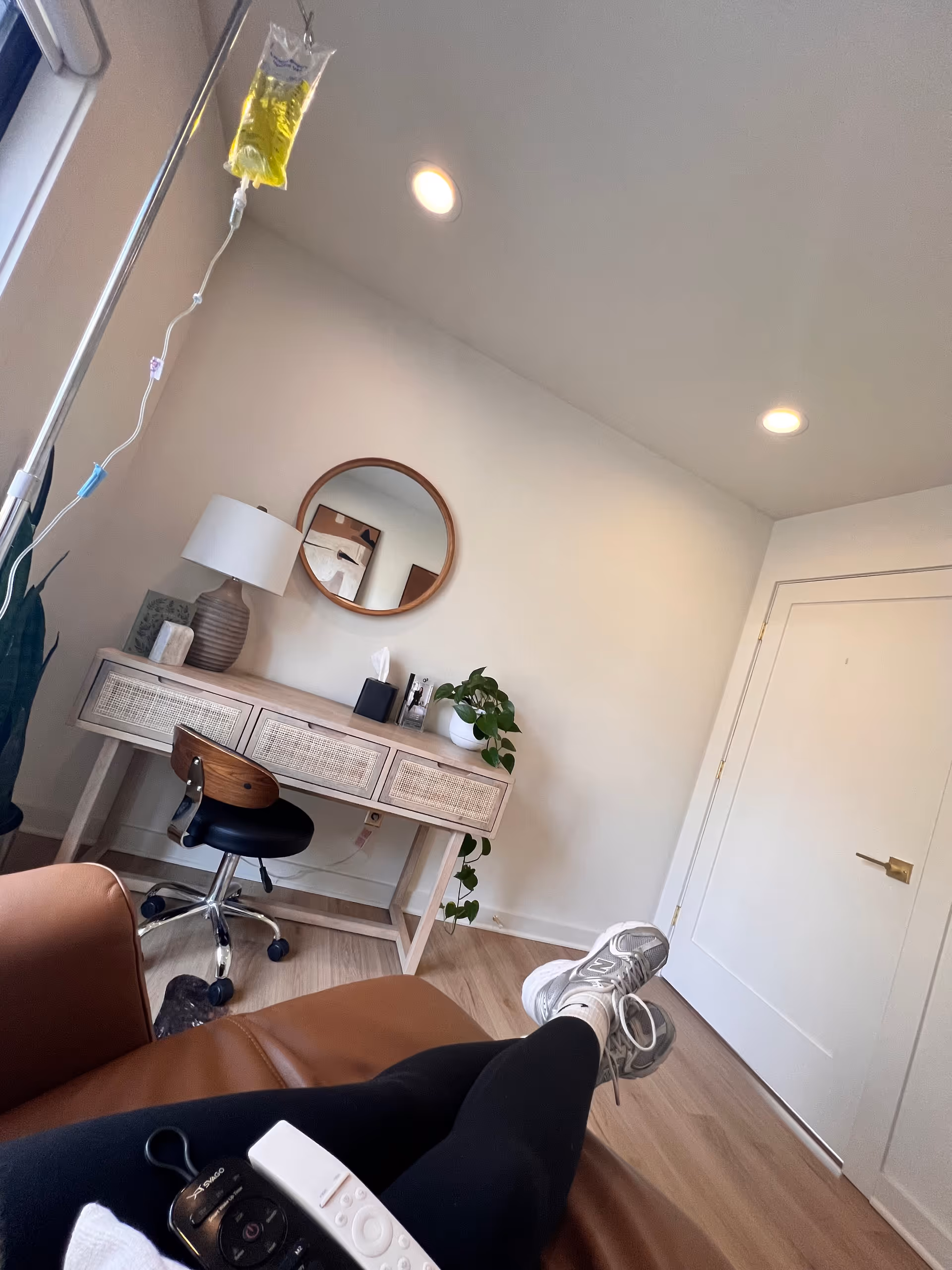 A wellness treatment room with IV drip, desk, mirror, chair, and a plant.
