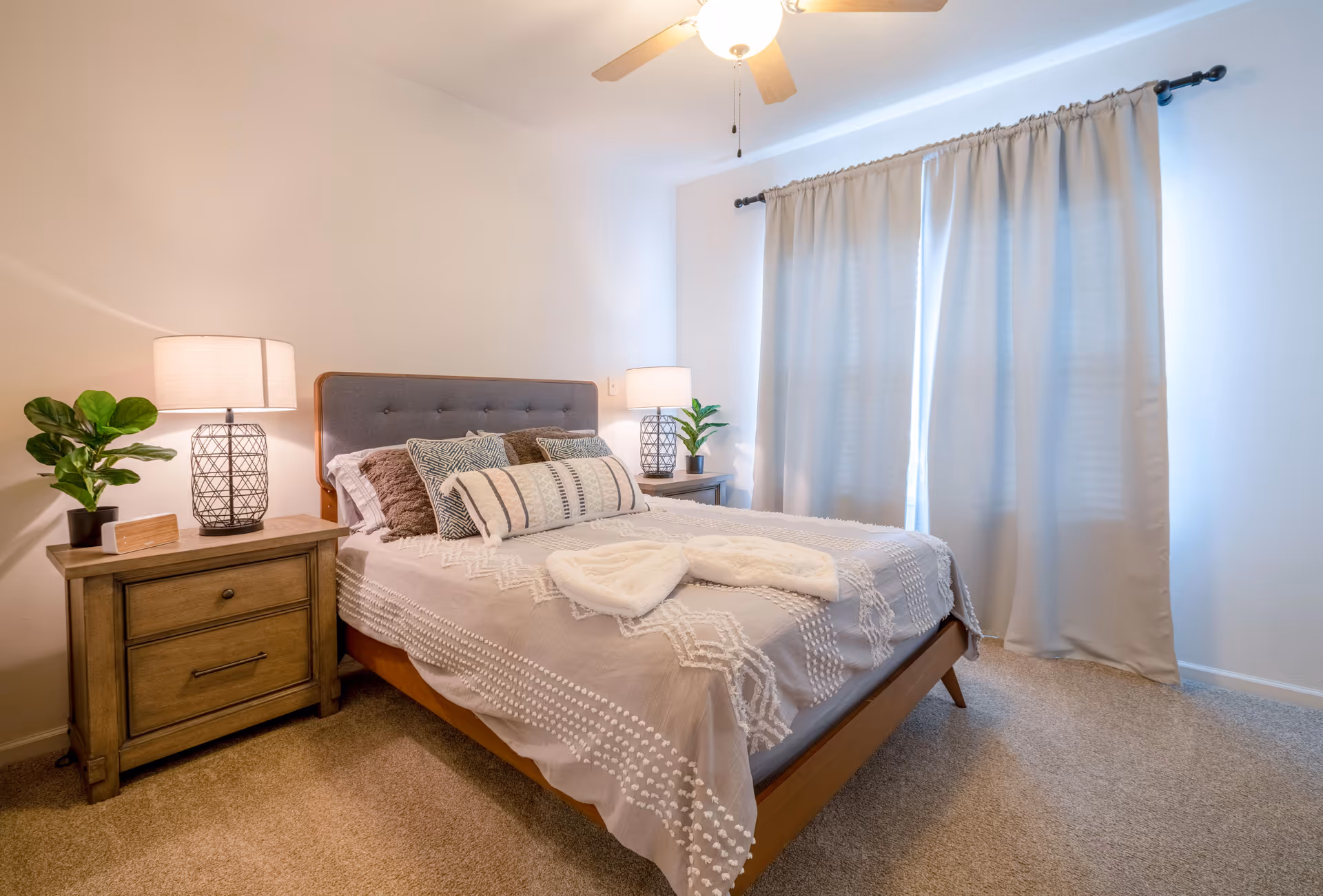 A neatly made bedroom with a bed, nightstands, lamps, plants, and curtains.