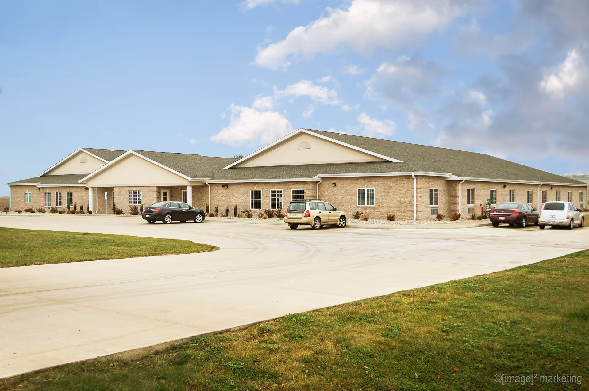 Exterior view of a single-story brick building with a pitched roof, several windows, and a few cars parked in front on a concrete driveway under a partly cloudy sky.