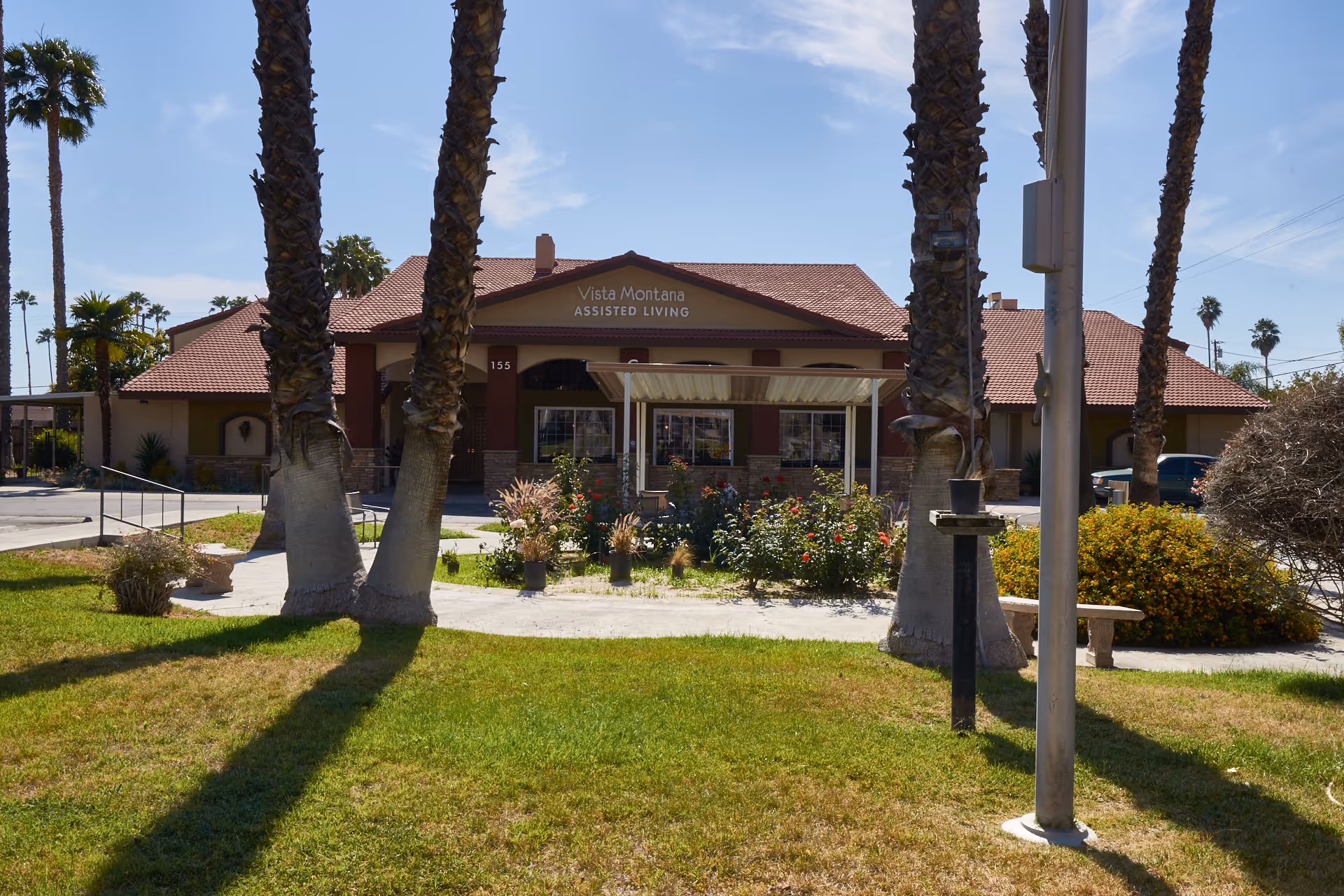 Front exterior view of Vista Montana Assisted Living facility with a red-tiled roof, surrounded by palm trees and landscaped greenery under a clear blue sky.