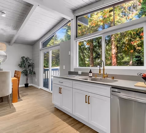 Bright kitchen interior with white cabinets, gold handles, a stainless steel dishwasher, and a sink under large windows showing green trees outside. Adjacent dining area with wooden table and chairs, light wood flooring, and a potted plant near sliding glass doors leading to an outdoor deck.