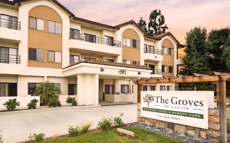 Front exterior of a three-story assisted living building with a driveway and a sign reading 'The Groves of Tustin'.