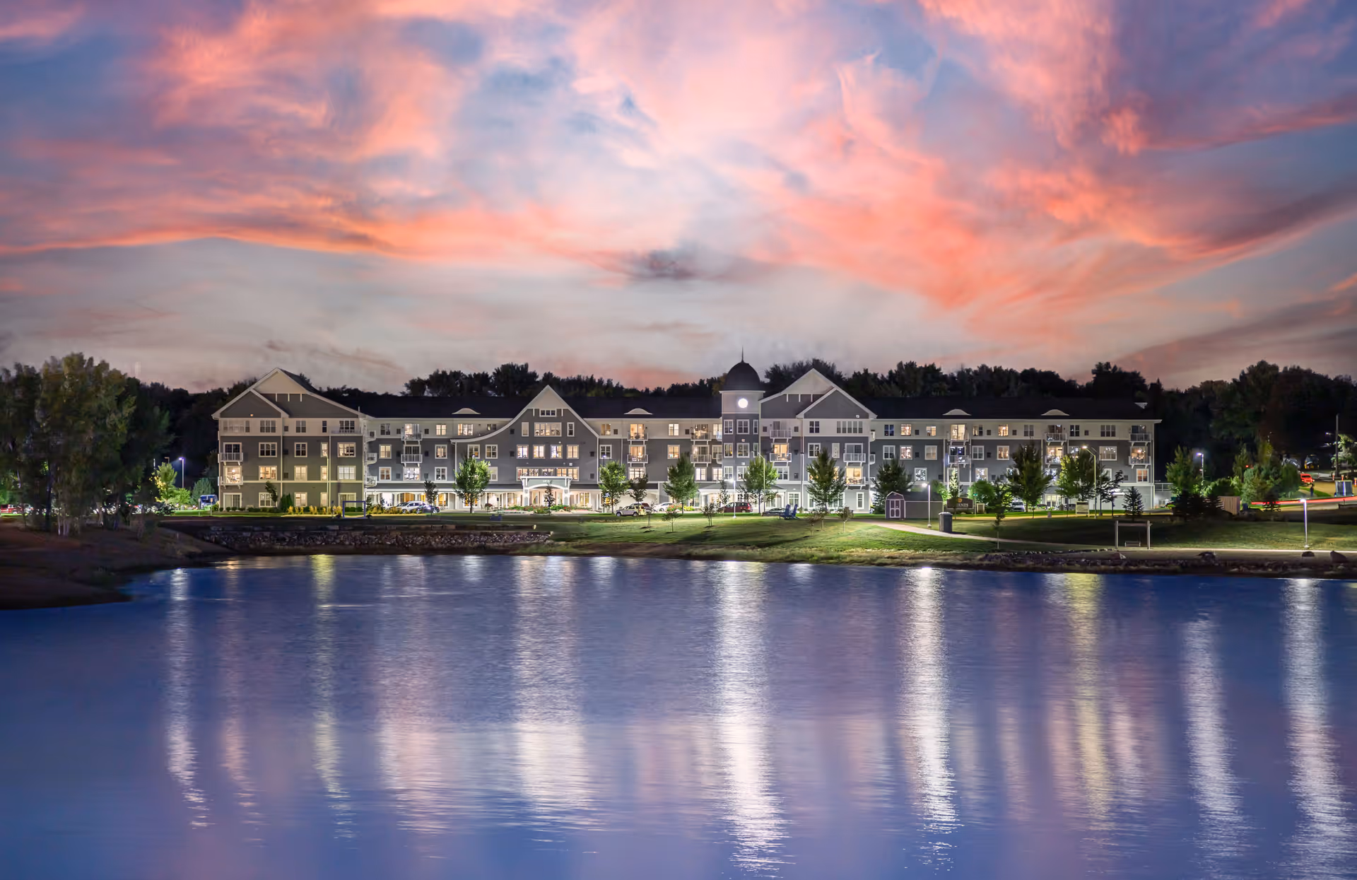 A long multi-story senior living building lit at dusk with a colorful pink sky reflected in the calm lake in front.