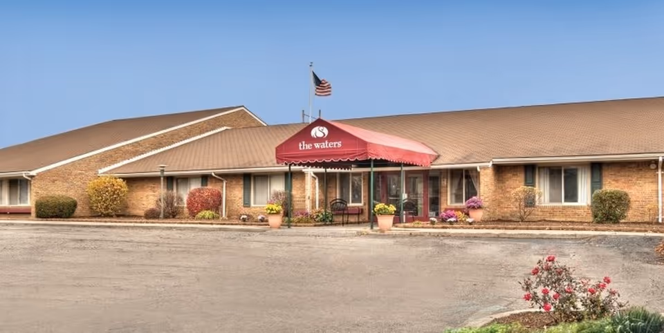 Front entrance of a single-story brick senior living facility with a red awning reading "the waters" and an American flag above.