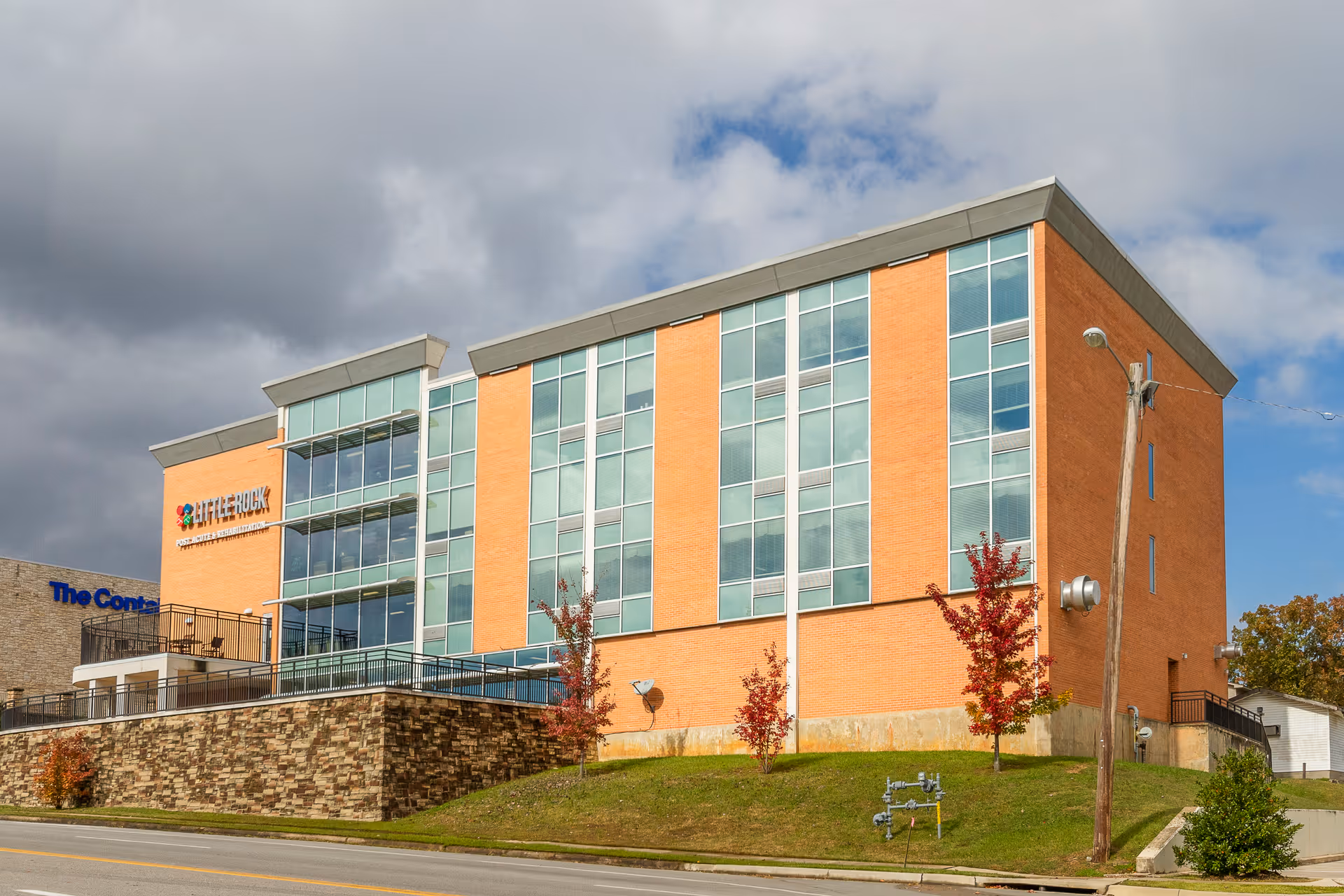 Brick multi-story nursing facility building with large vertical windows on a grassy slope under a cloudy sky.