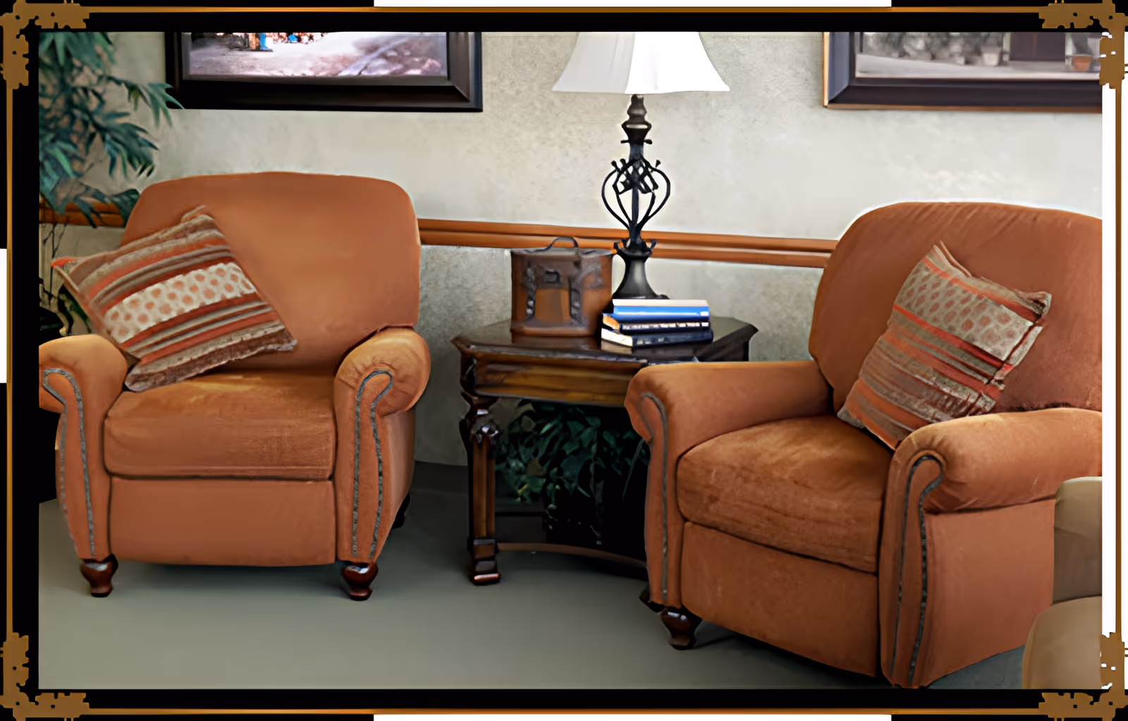 Two orange upholstered armchairs with striped cushions are positioned on either side of a wooden side table. The table holds a decorative lamp and a small stack of books. The background includes framed artwork and a plant partially visible on the left side.