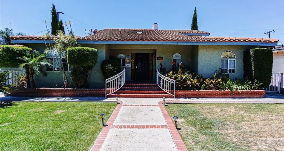 Front exterior view of a single-story house with a tiled roof, a concrete walkway bordered by red bricks leading to the entrance, and landscaped greenery including trimmed bushes and palm trees on either side.