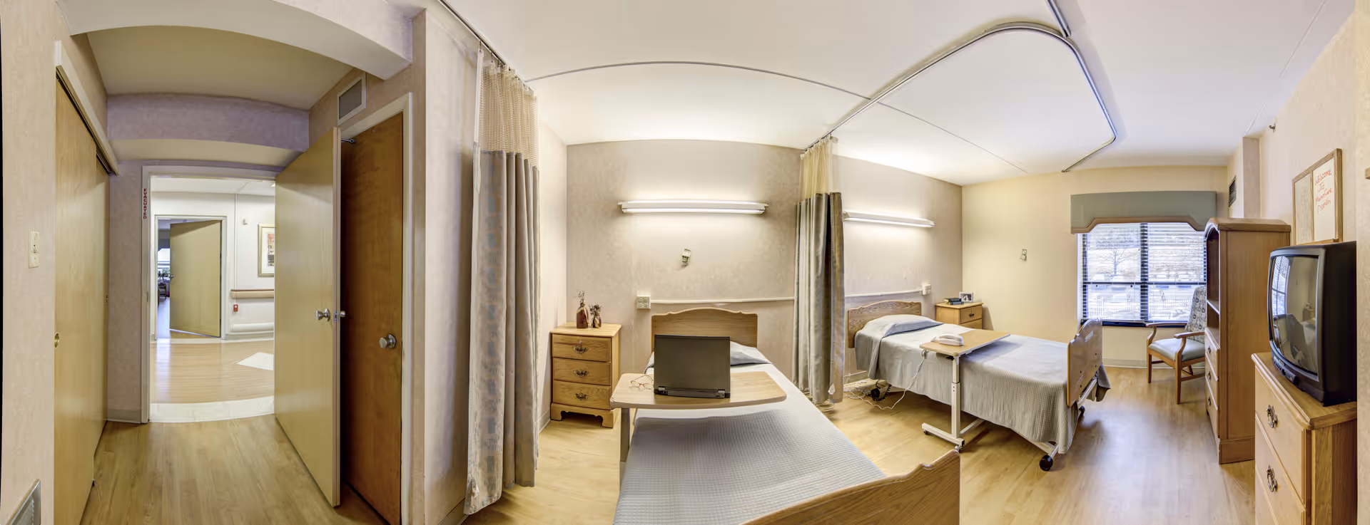 Panoramic view of a two-bed patient room with curtains, desks, and a window.