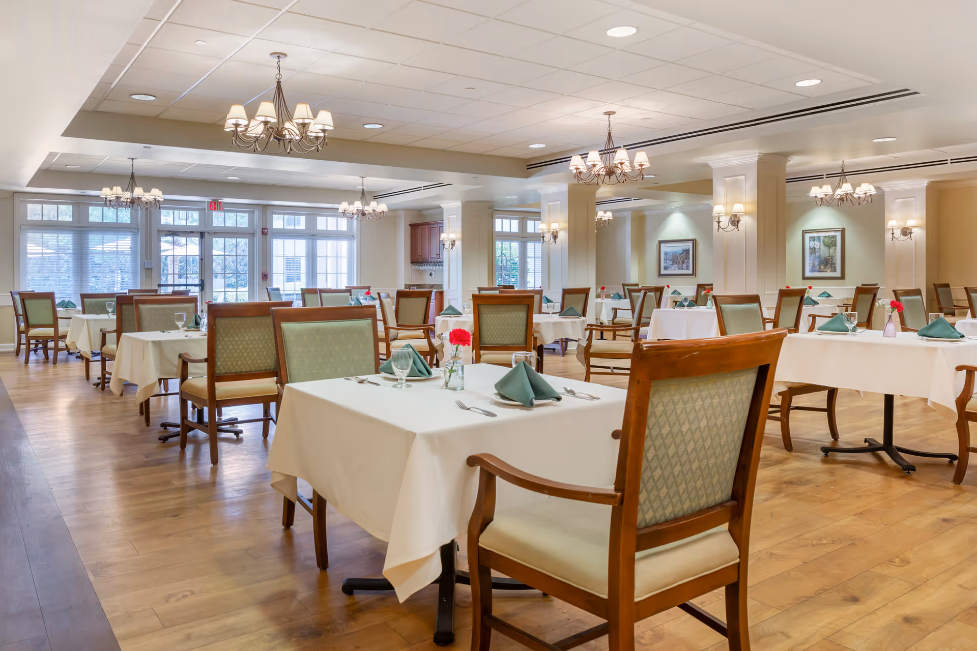 Bright dining room with white tablecloths, green napkins, and chandeliers in a senior residence.