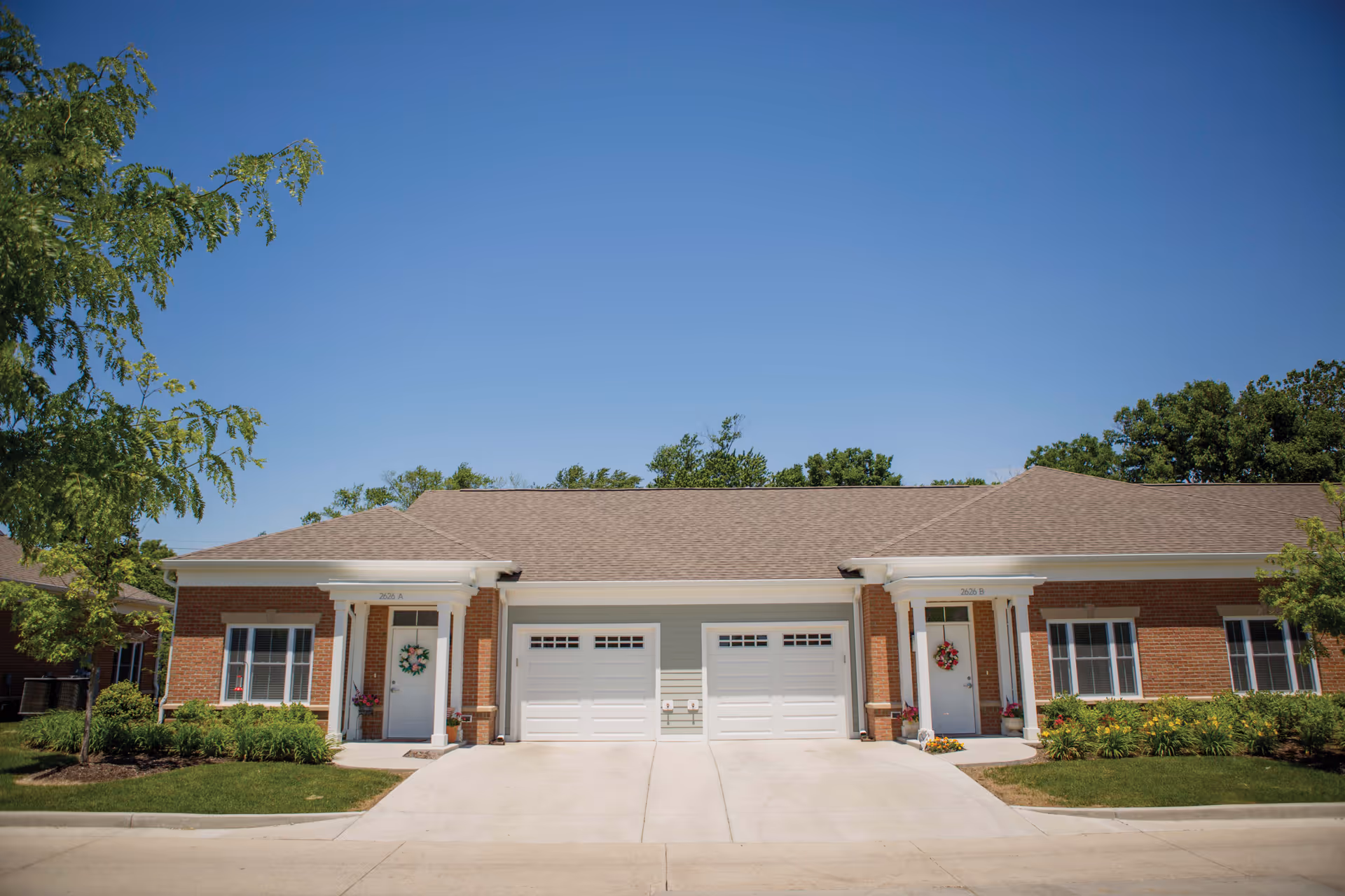 Front view of a single-story duplex with two garage doors, brick facades, wreath-adorned entry doors, and landscaping under a clear blue sky.