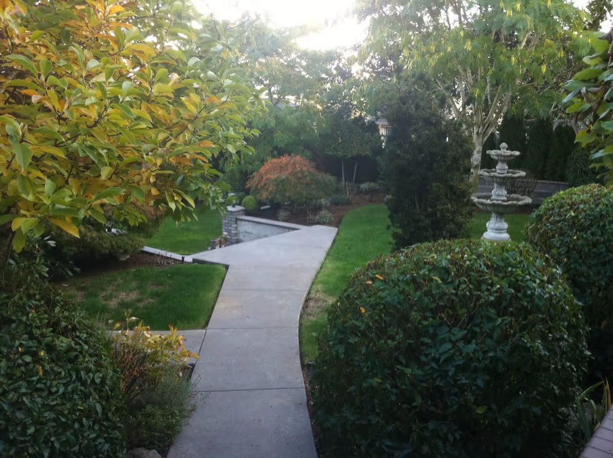 A paved walkway winding through a well-maintained garden with green grass, various bushes, trees, and a decorative three-tiered stone fountain on the right side.