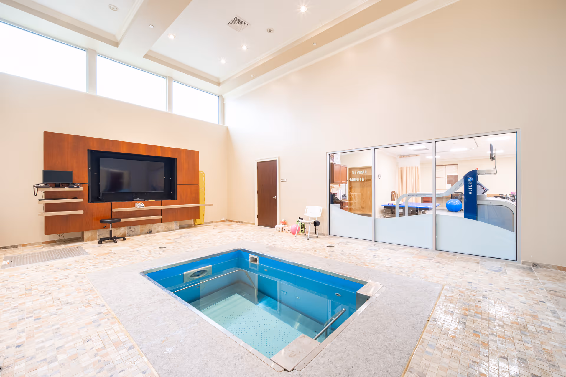 Bright indoor therapy room featuring a small sunken exercise pool, a wall-mounted TV on a wood panel, and a glass-fronted therapy area with equipment.