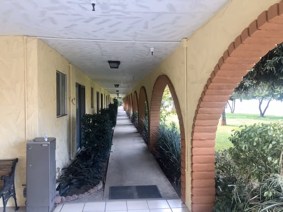 Covered walkway with a row of arched brick openings beside landscaped lawn and apartment doors.