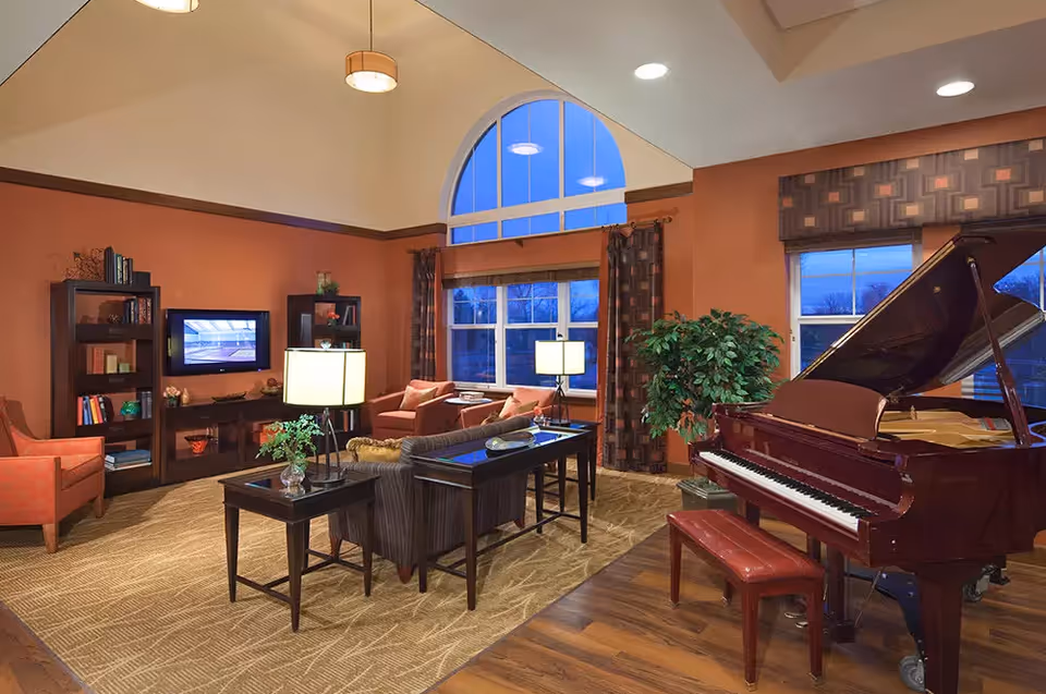 A cozy living room area in an assisted living facility featuring a grand piano with a bench, comfortable seating including armchairs and a sofa, a coffee table with a lamp and plant, bookshelves with books and decorative items, large windows with curtains, and warm lighting from ceiling fixtures and table lamps.