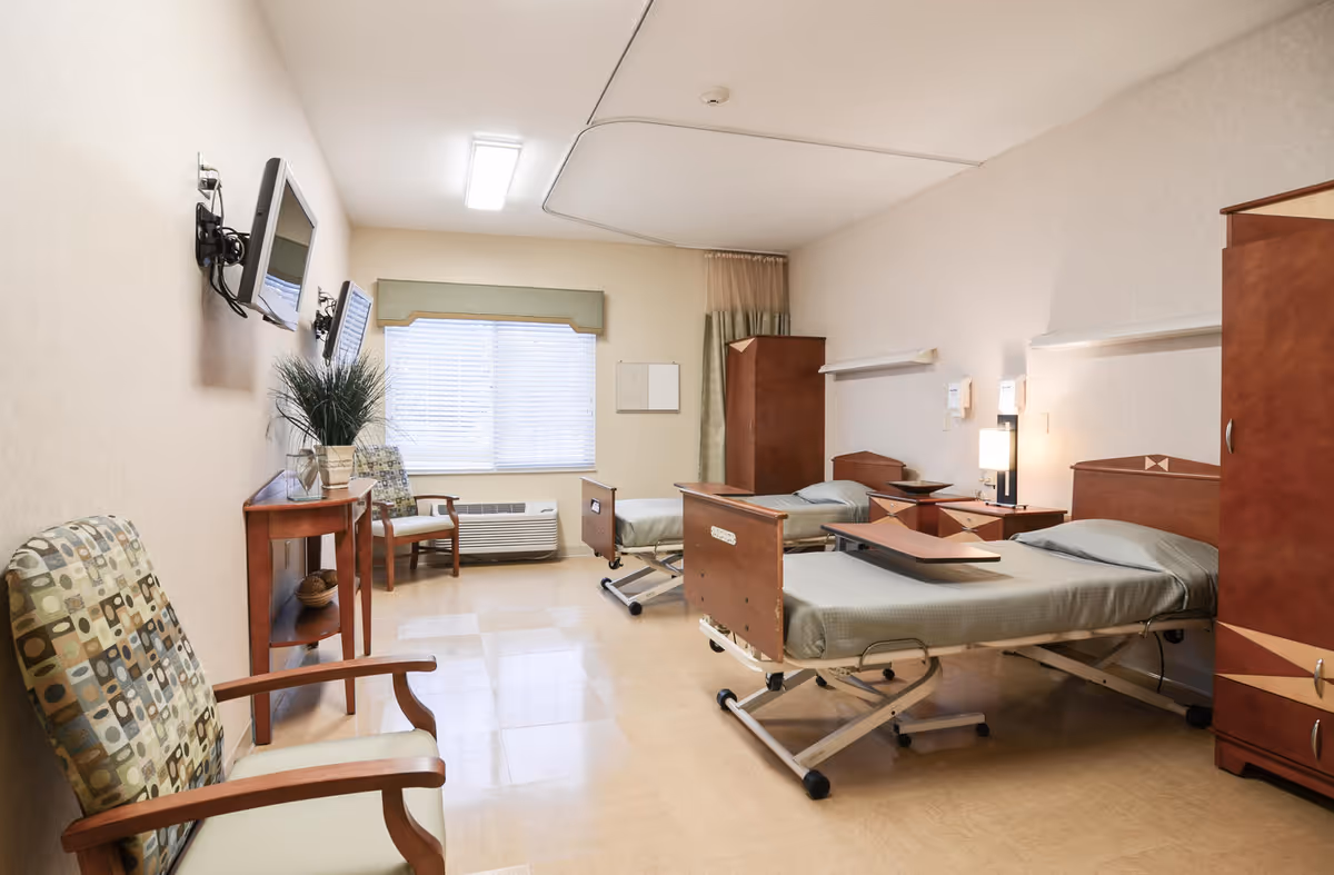 Shared senior living room with multiple beds, chairs, and a wall-mounted TV.