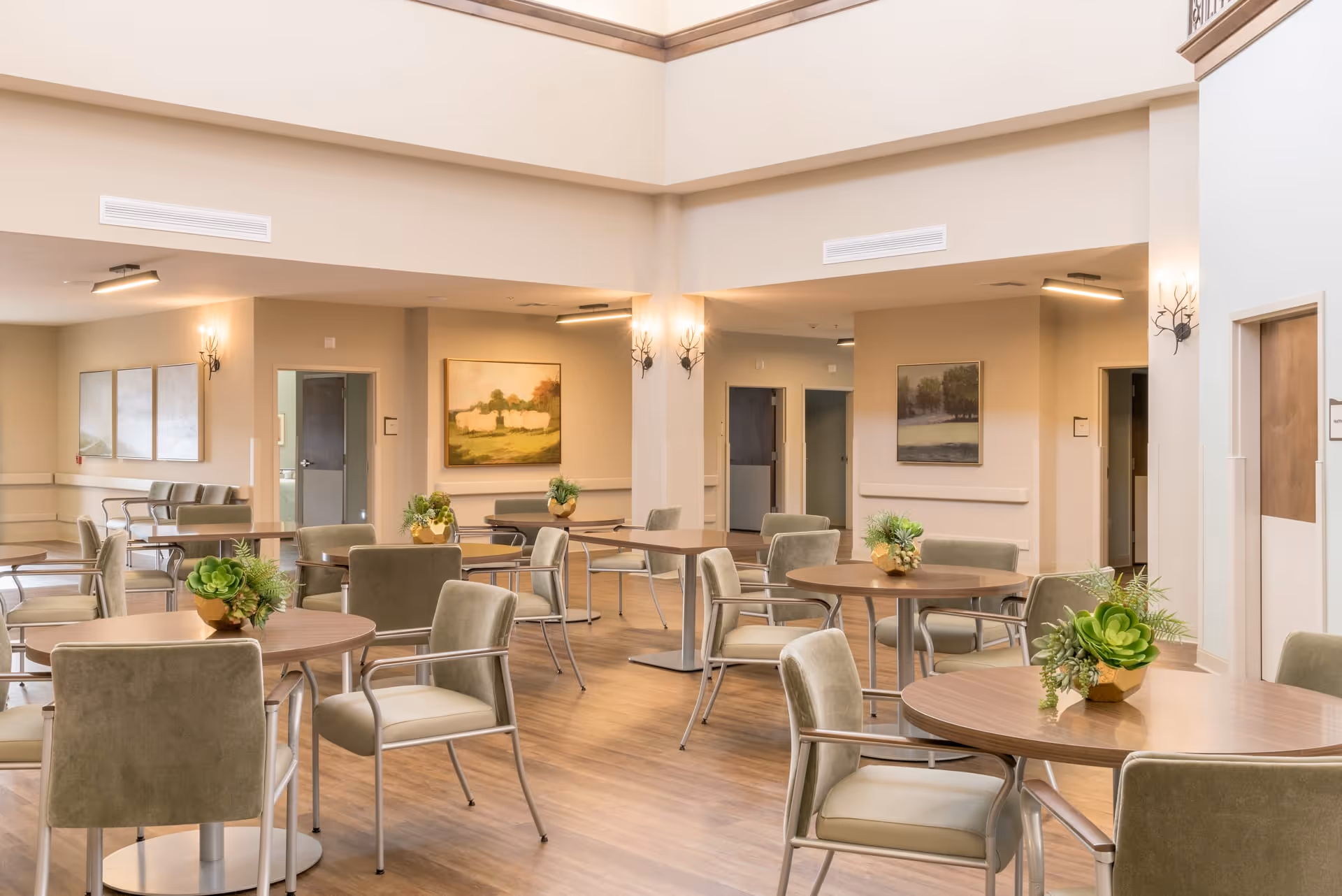 A spacious and well-lit dining area with round wooden tables and cushioned chairs arranged neatly. Each table has a small green plant centerpiece. The room has light-colored walls adorned with landscape paintings and soft wall-mounted lighting fixtures. The floor is wooden, and there are multiple doorways leading to other rooms.