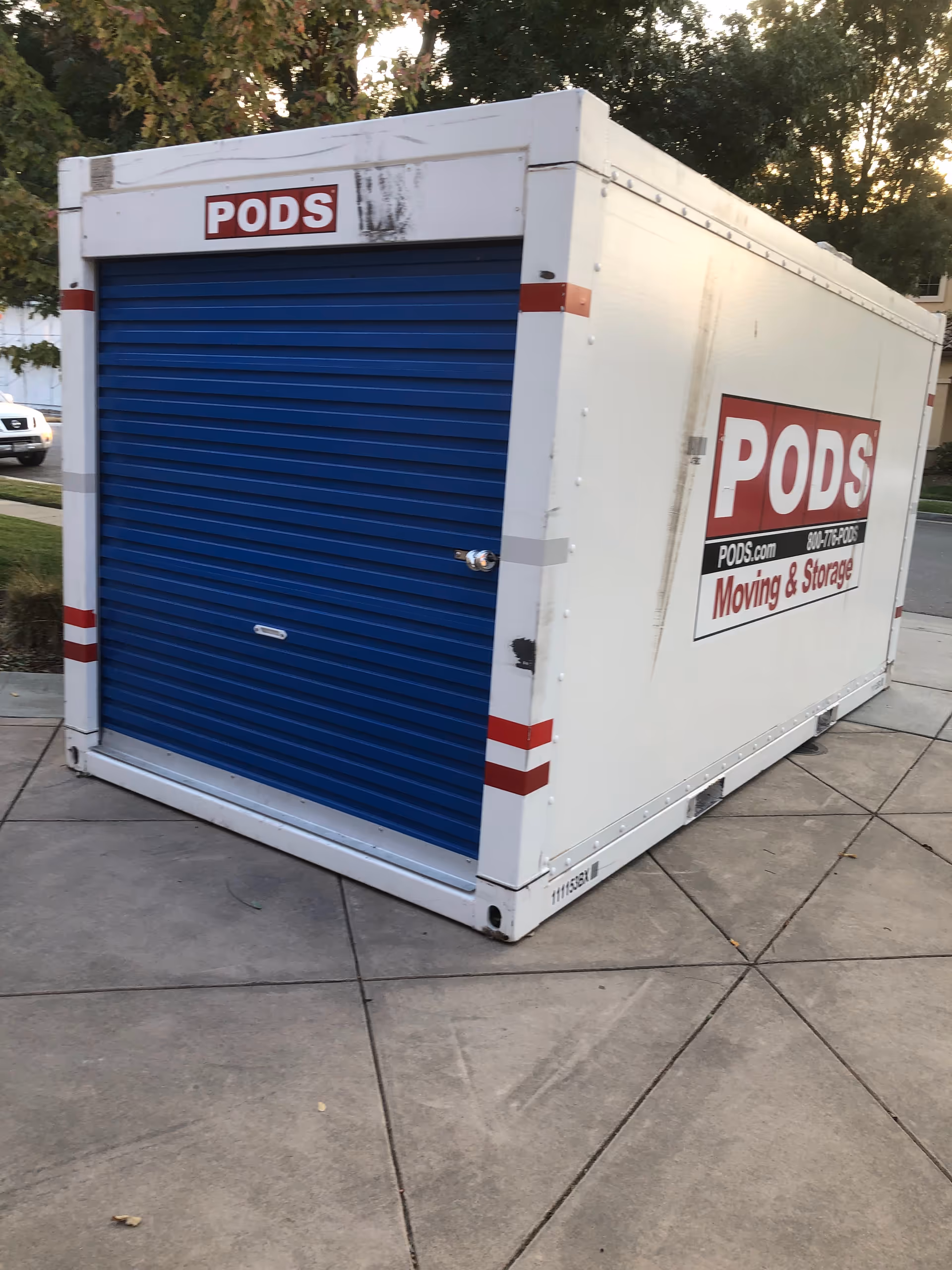 A white PODS storage container with a blue roll-up door on a paved outdoor area.