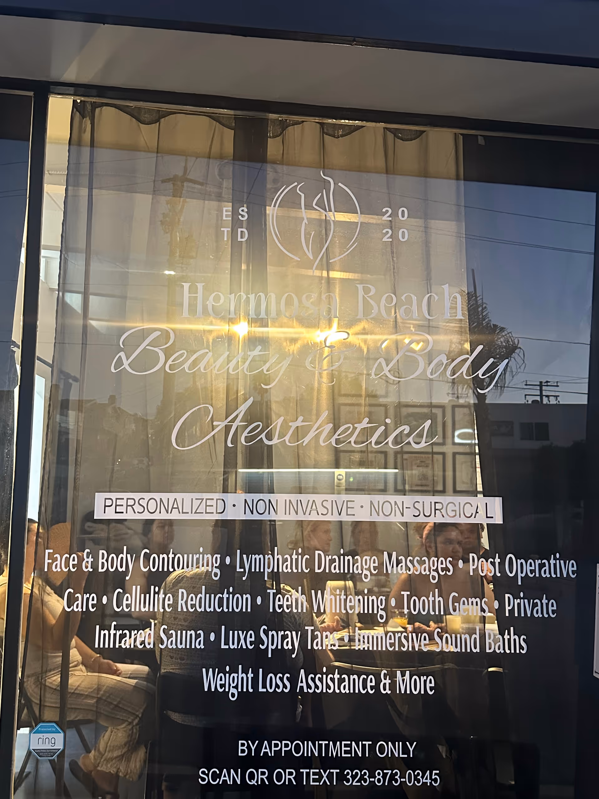 Glass storefront window with Hermosa Beach Beauty & Body Aesthetics signage and customers inside.