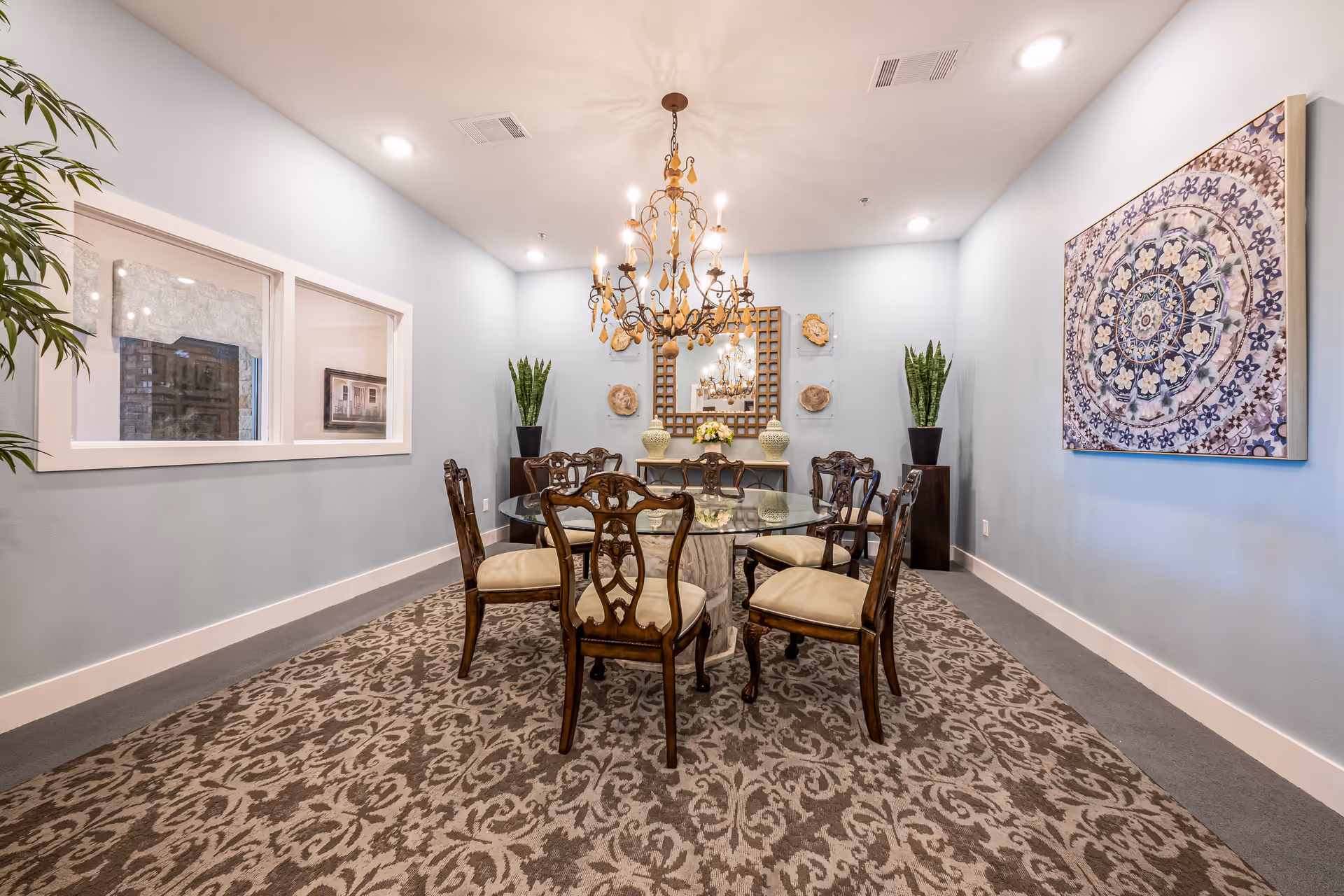 A formal dining room with a round glass table, chairs, chandelier, and wall art.