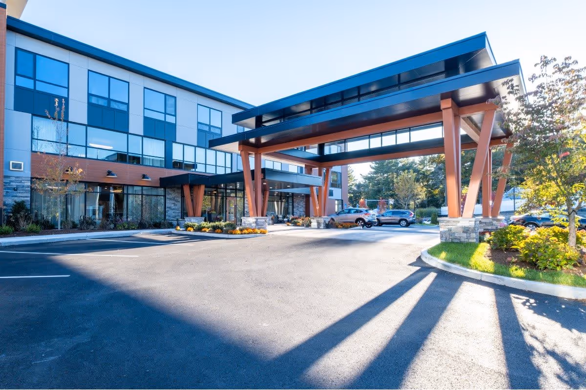 Exterior view of Benchmark at Hanover senior living facility showing the main entrance with a large covered drop-off area supported by wooden beams, a parking lot with several cars, and landscaped greenery around the building.