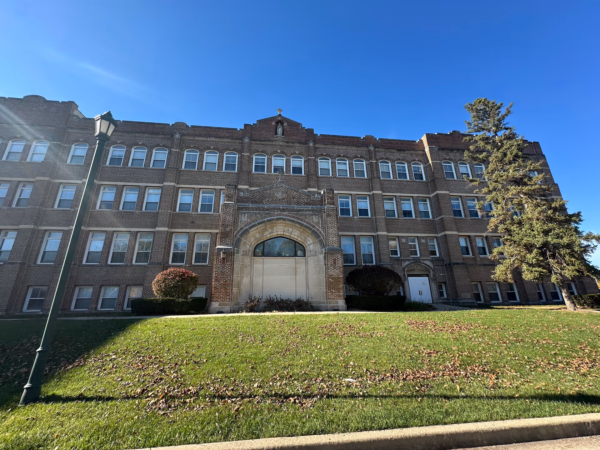 Front view of a large multi-story brick building with a central arched entrance, lawn, lamp post and trees under a clear blue sky.