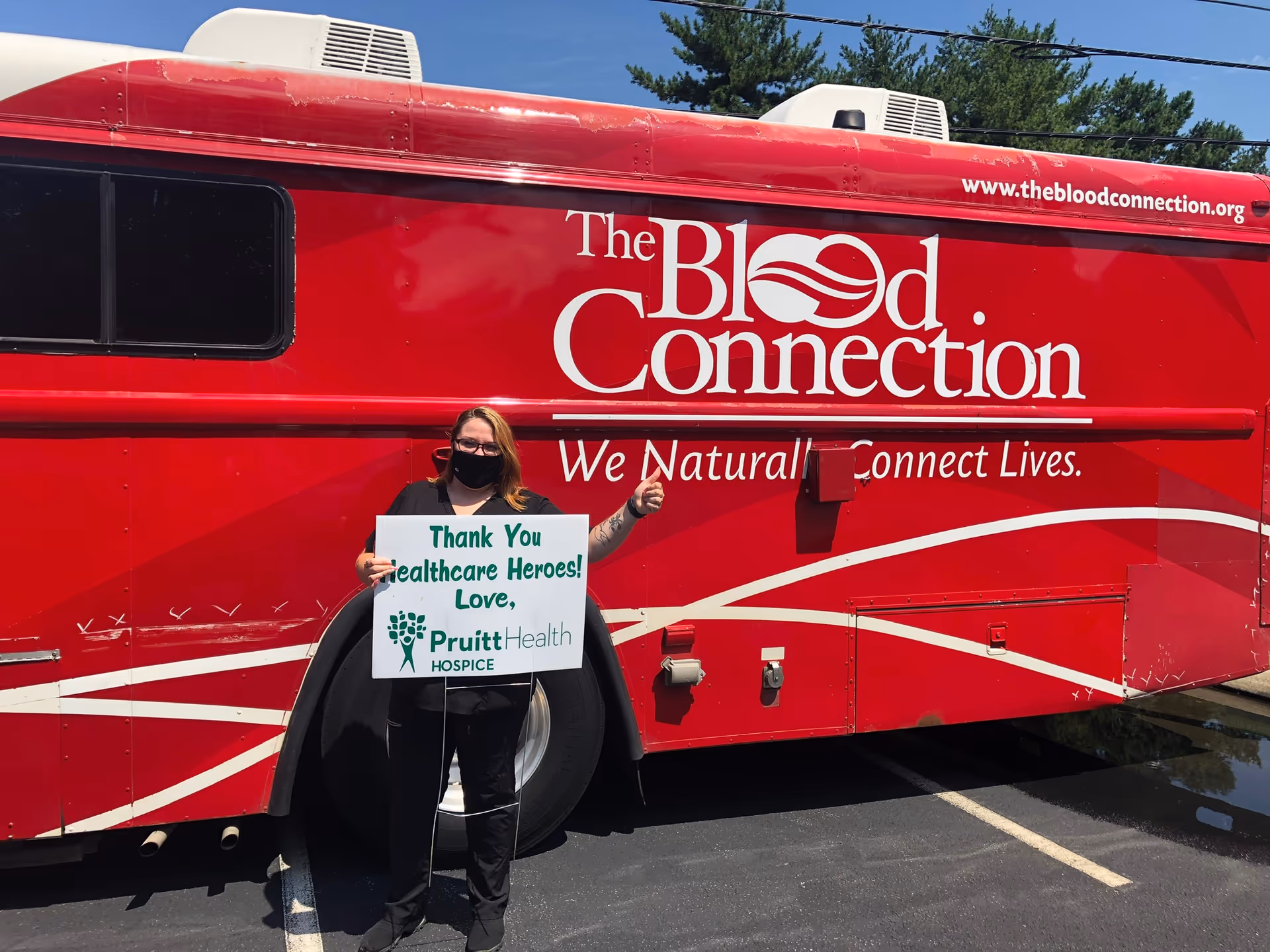A person stands beside a large red blood donation bus with a sign.
