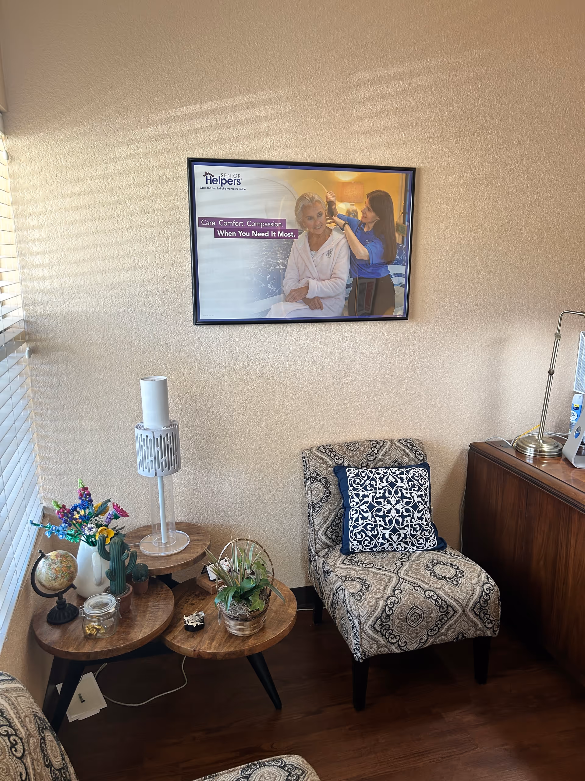 Cozy living-room corner with patterned chair, small tables, plants, and a framed wall poster.