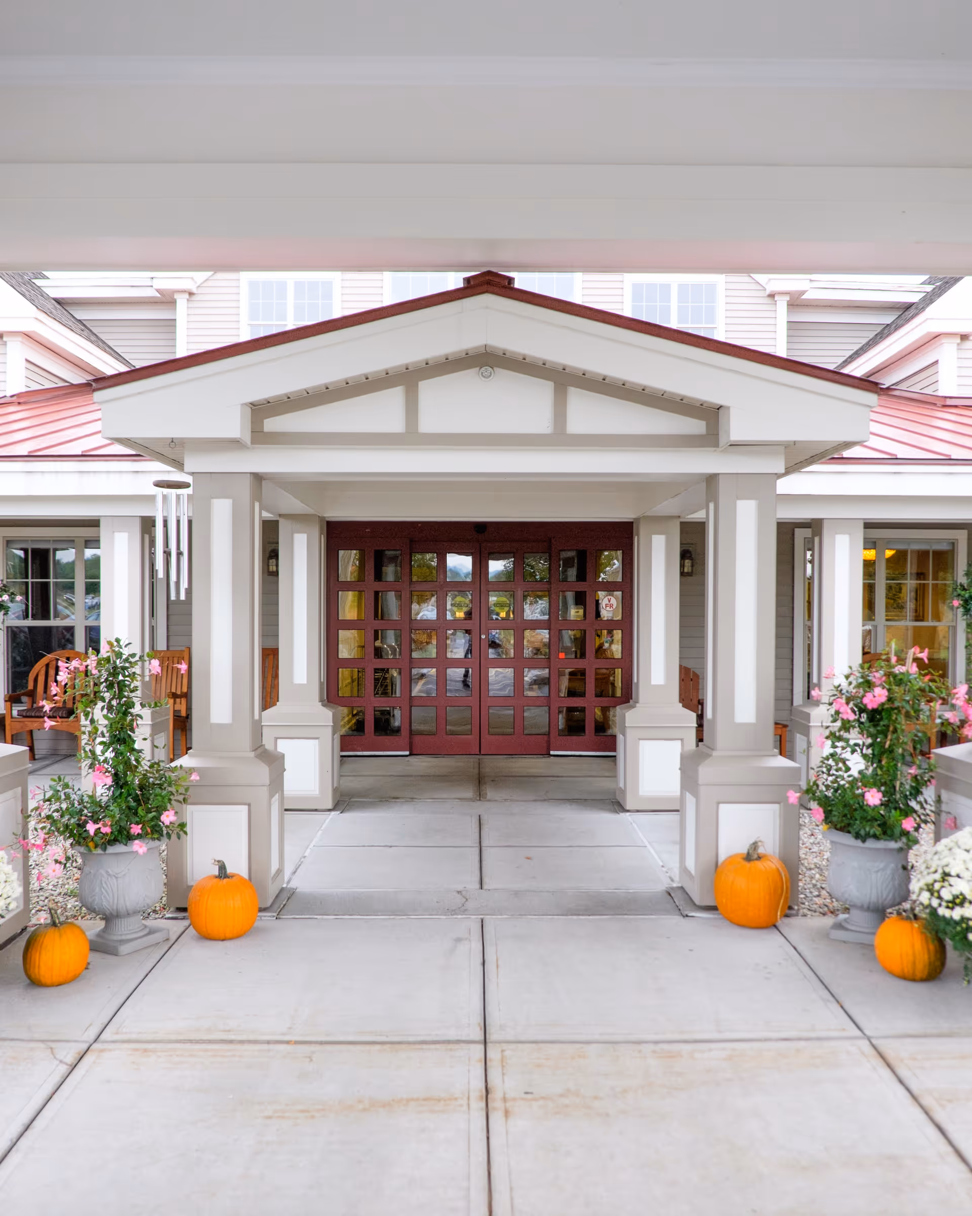 Entrance to a senior living facility with a covered walkway supported by columns. The entrance features double red doors with glass panes. There are pumpkins and flower pots with pink flowers on either side of the walkway, suggesting a fall or autumn season.