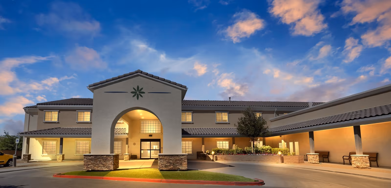 Exterior view of Inspirations Of Pahrump facility at dusk, showing a two-story building with a tiled roof, an arched entrance with stone accents, illuminated windows, and a landscaped area with benches and plants.