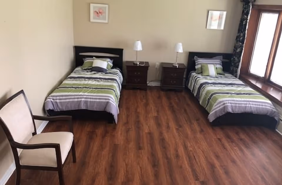 A shared bedroom with two twin beds, nightstands with lamps, a chair, wood floor, and windows.