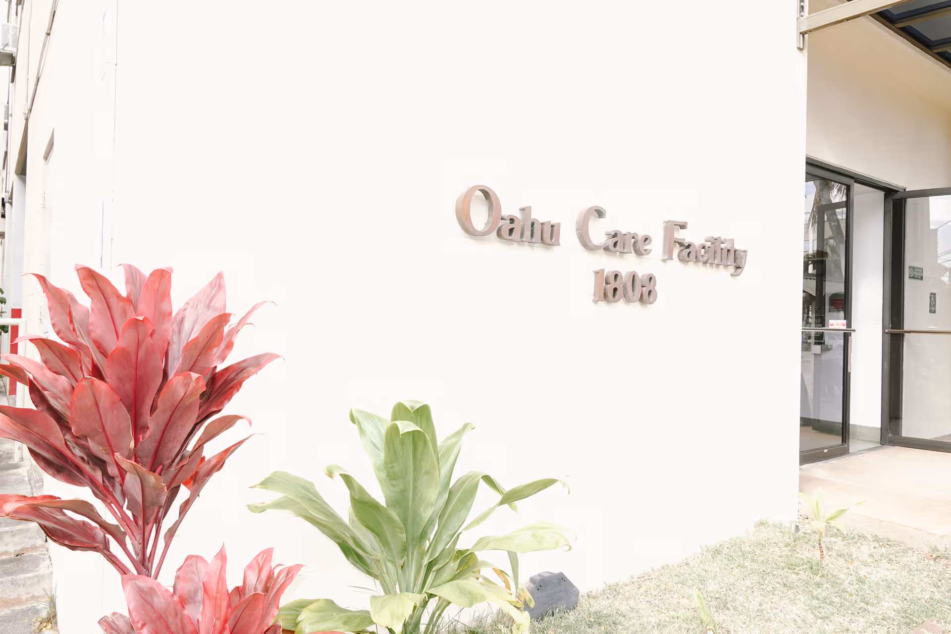 White building exterior with Oahu Care Facility sign and tropical plants near the entrance.