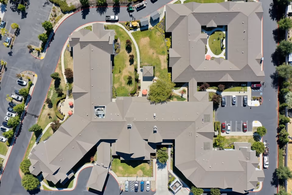 Aerial view of The Commons At Dallas Ranch facility showing multiple connected buildings with gray roofs, surrounding green lawns, pathways, parking lots with cars, and some trees and landscaping.