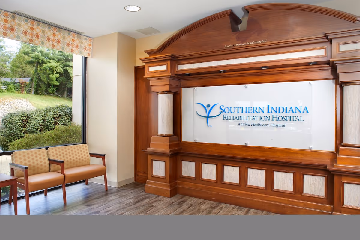 Interior view of the Southern Indiana Rehabilitation Hospital lobby area featuring a large wooden decorative wall panel with the hospital's name and logo. To the left, there are two cushioned chairs next to a large window showing greenery outside.