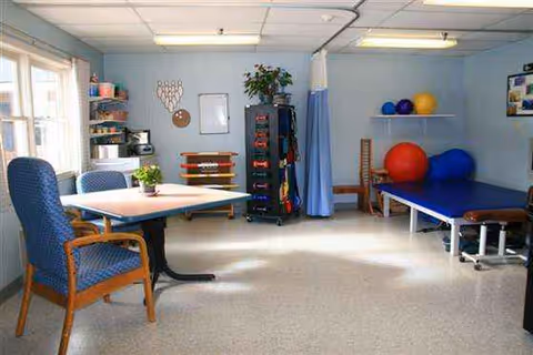 Rehabilitation room with a table, chairs, shelves, exercise equipment, and a treatment bed.