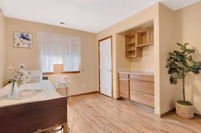 A simple beige senior living bedroom with bed, nightstand, dresser, window, and potted plant.