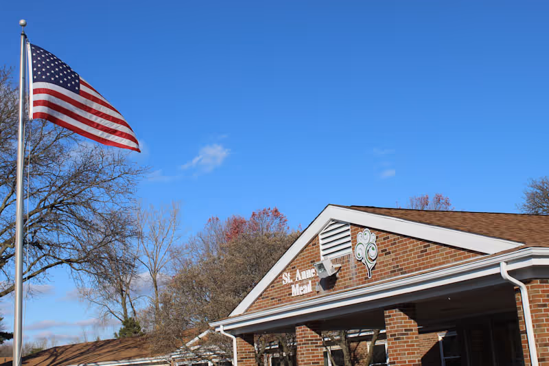 Front exterior of the St. Anne's Mead brick building with an American flag flying on a flagpole under a clear blue sky.