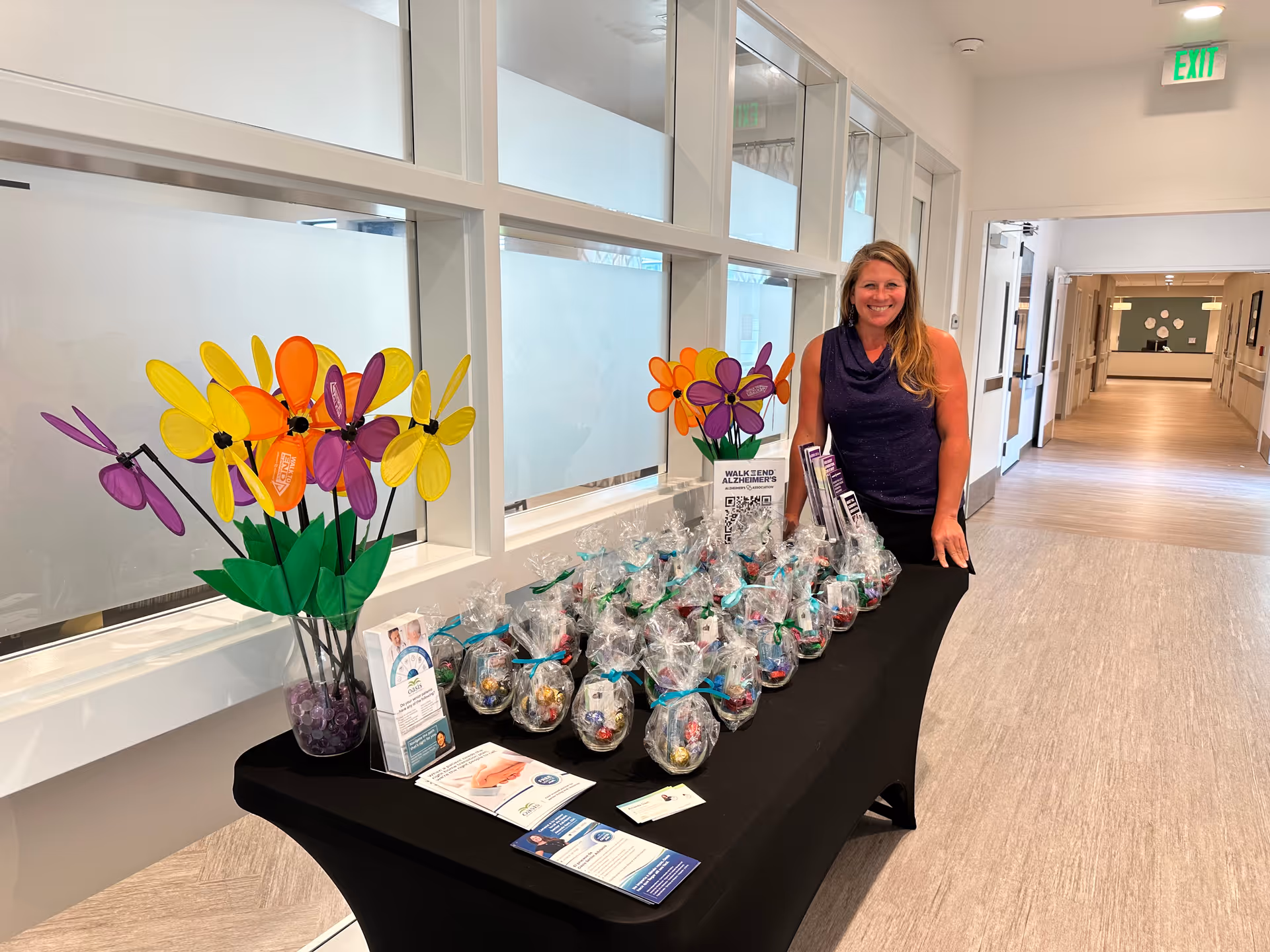 Smiling staff member stands beside a gift display on a corridor table in a senior facility.