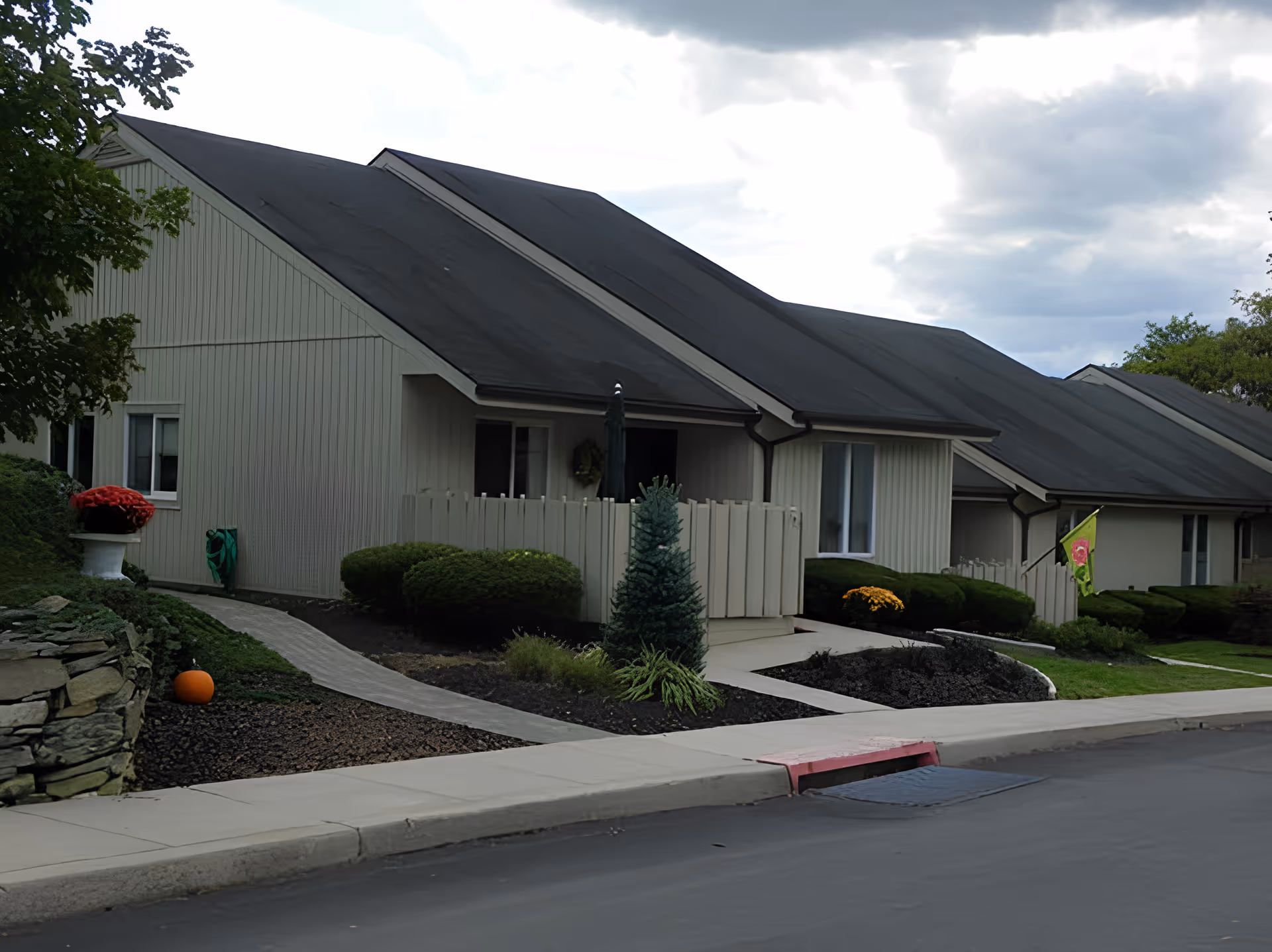 Front exterior of single-story beige townhouse-style units with dark roofs, small fenced patios, and landscaped walkways.