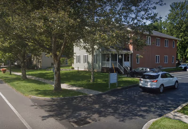 Two-story building with a front porch, trees, and parked cars along a street.