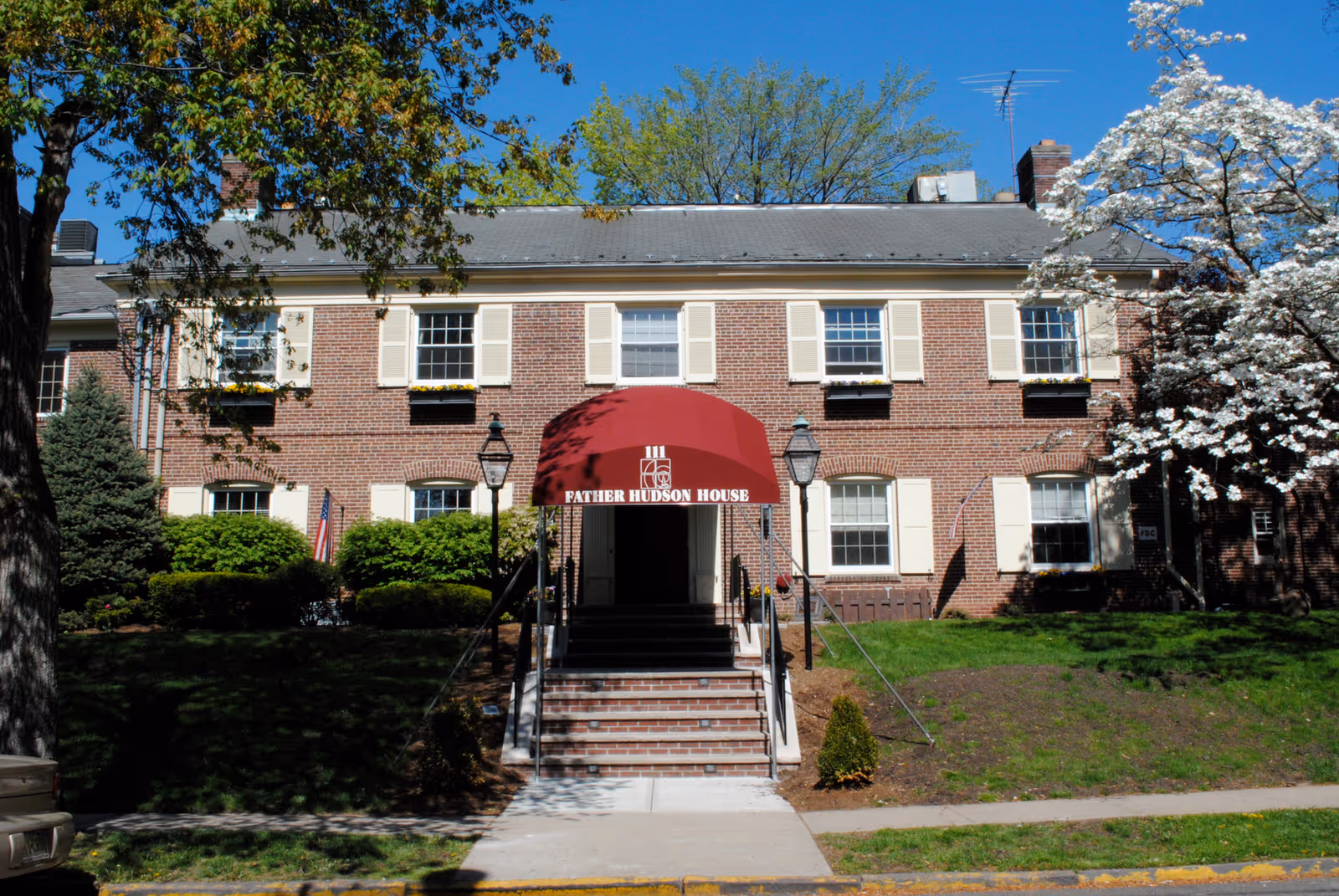 Two-story brick building with a red entrance awning labeled 'Father Hudson House', front steps, and surrounding trees and lawn.