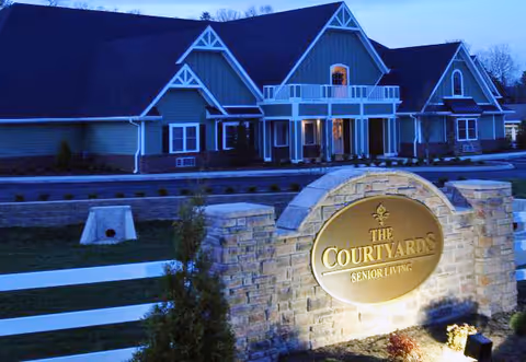 Exterior view of The Courtyards Senior Living facility at dusk, showing a large building with multiple peaked roofs and a well-lit stone sign in the foreground displaying the facility's name.