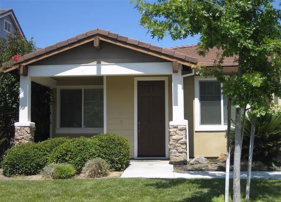 Front exterior of a single-story house with a covered porch, brown front door, windows, and trimmed shrubs and lawn.