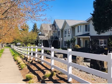A two-story residential building with white siding and dark brick accents, surrounded by trees and landscaping. A white wooden fence runs alongside a sidewalk, casting shadows on the ground under a clear blue sky.