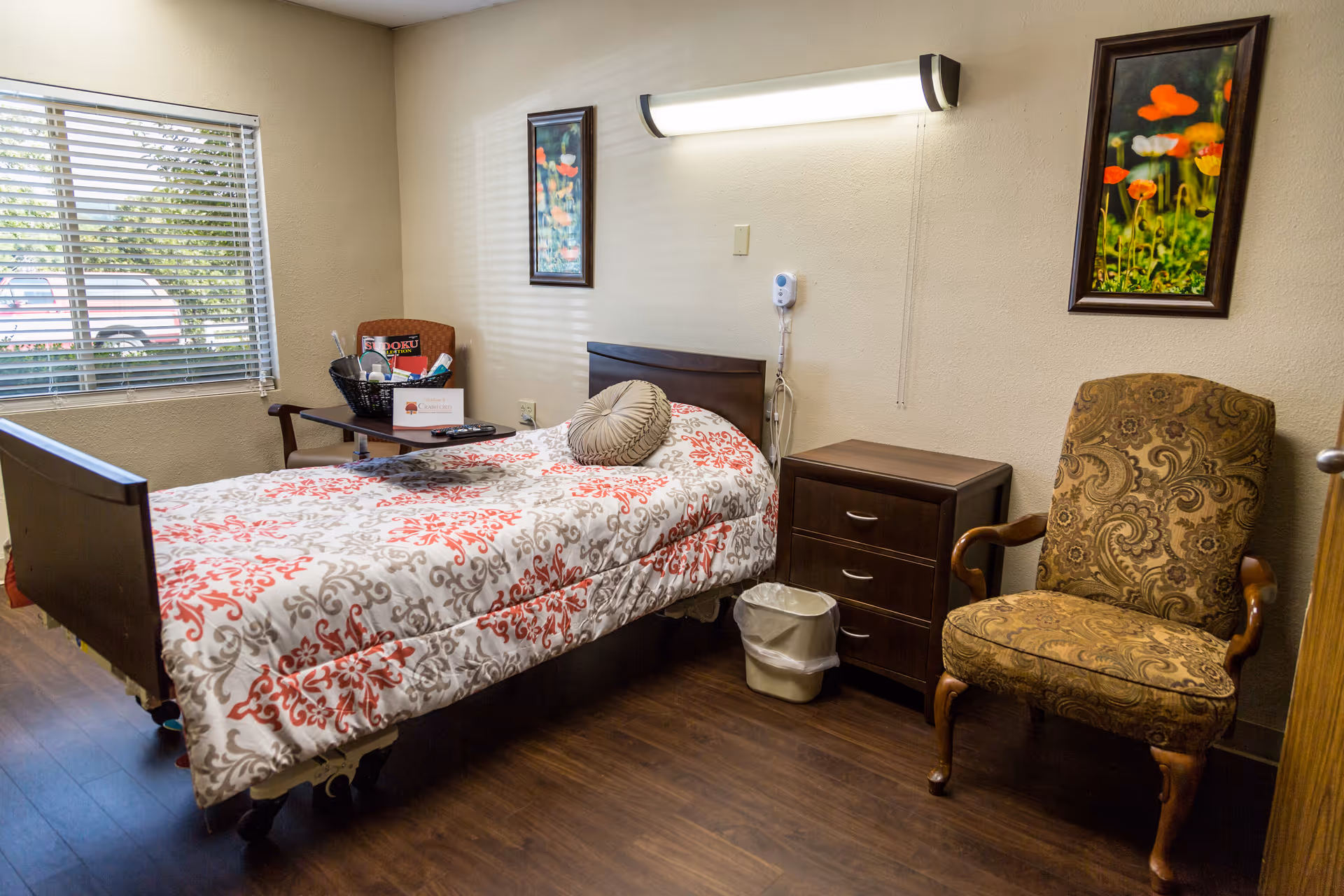 A hospital-style bedroom with a single bed, nightstand, window, and paisley chair.