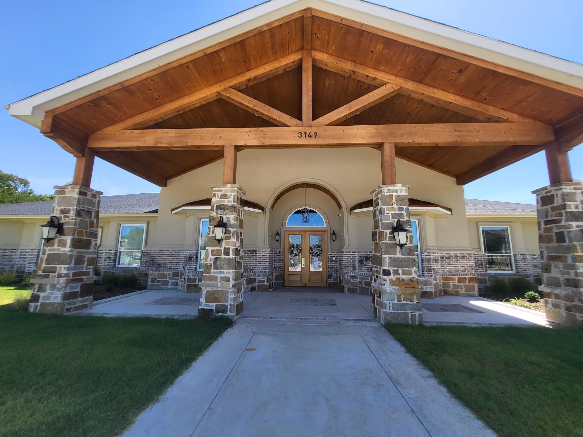 Front exterior view of a building with a large wooden covered entrance supported by stone pillars. The building has a combination of stone and brick facade with a double wooden door entrance under an arched window. The address number 3149 is visible above the entrance.