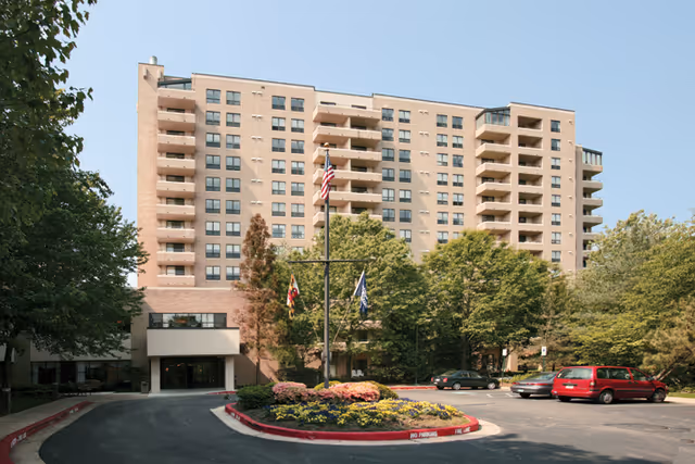 A multi-story beige apartment building with balconies, flagpoles, and a circular driveway entrance surrounded by trees and landscaping.