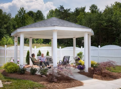 Two elderly people sit and chat under a white gazebo in a fenced garden.