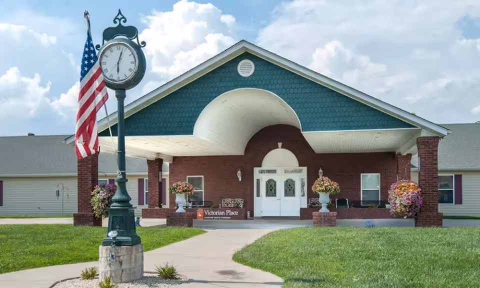Front entrance of a single-story senior living building with a covered portico, ornamental clock and an American flag on the lawn.